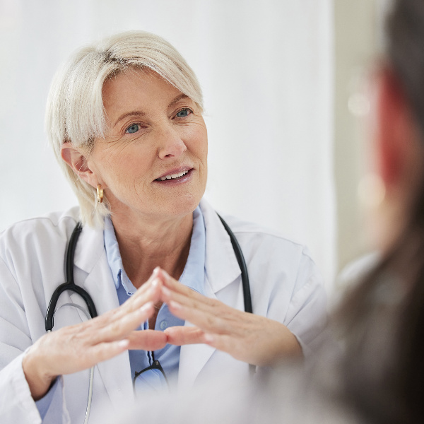 Ärztin im weißen Arztkittel mit Stethoskop stützt ihre Ellbogen auf einem Tisch und ist im Gespräch mit einer Patientin.