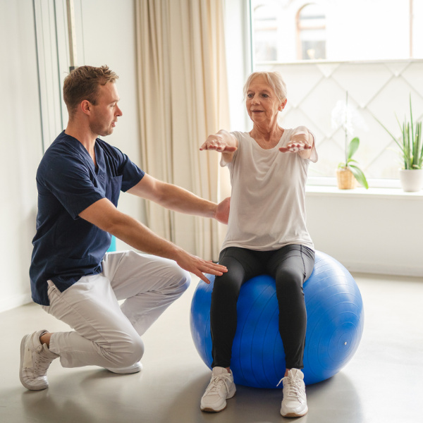 Ältere Frau macht Übungen mit Physiotherapeuten auf einem blauen Gymnastikball und hält dabei ihre Arme nach vorne ausgestreckt. Ältere Frau macht Übungen mit Physiotherapeuten auf einem blauen Gymnastikball und hält dabei ihre Arme nach vorne ausgestreckt.