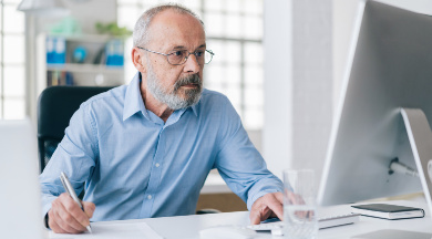 Ein älterer Mann im blauen Hemd und Brille arbeitet am Schreibtisch und blickt auf einen Computer-Bildschirm. Ein älterer Mann im blauen Hemd und Brille arbeitet am Schreibtisch und blickt auf einen Computer-Bildschirm.