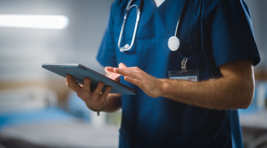Ein Arzt in blauer Uniform mit Stethoskop, der auf einem Tablet arbeitet. Der verschwommene Hintergrund zeigt ein Patientenzimmer.