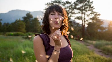 Eine Frau mit schulterlangen braunen Haaren steht auf einer Wiese und pustet auf eine Pusteblume.