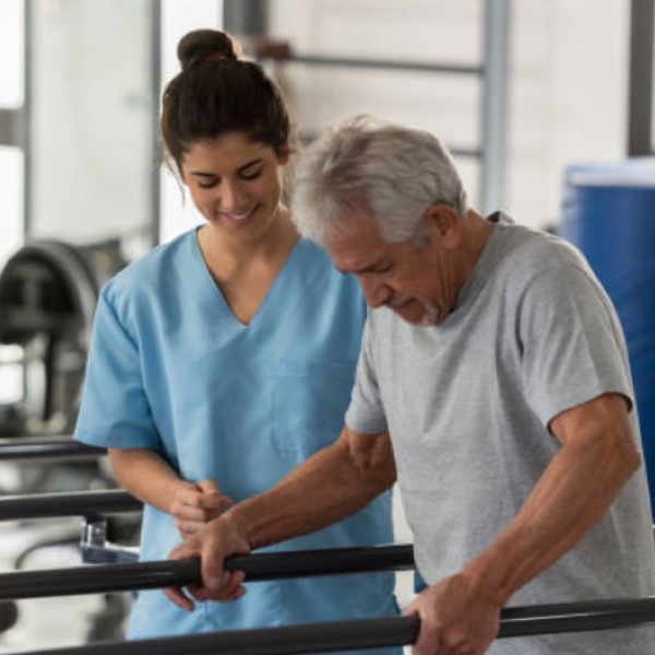 Älterer Mann wird von einer Physiotherapeutin in blauer Arbeitskleidung bei der Gehrehabilitation unterstützt. Älterer Mann wird von einer Physiotherapeutin in blauer Arbeitskleidung bei der Gehrehabilitation unterstützt.