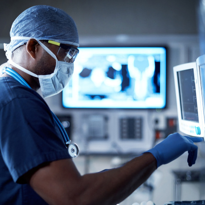 Ein Arzt mit Stethoskop, steriler Kleidung und Handschuhen arbeitet im OP-Saal am Monitor. Ein Arzt mit Stethoskop, steriler Kleidung und Handschuhen arbeitet im OP-Saal am Monitor.