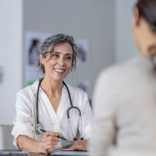 Ärztin mit Stethoskop lächelt und führt ein medizinisches Beratungsgespräch mit einer Patientin, die im Vordergrund sitzt. Ärztin mit Stethoskop lächelt und führt ein medizinisches Beratungsgespräch mit einer Patientin, die im Vordergrund sitzt.