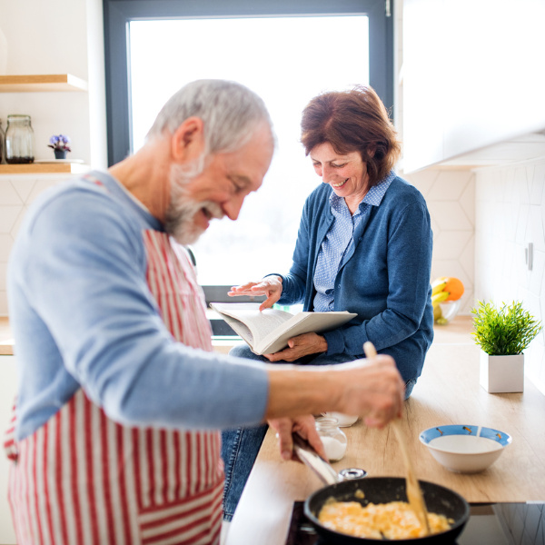 Ein älterer Mann mit gestreifter Schürze bereitet Essen in einer Pfanne zu, während die Frau auf der Küchenzeile sitzt und lächelnd durch ein Buch blättert. Ein älterer Mann mit gestreifter Schürze bereitet Essen in einer Pfanne zu, während die Frau auf der Küchenzeile sitzt und lächelnd durch ein Buch blättert.