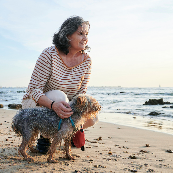 Eine ältere Frau hockt bei ihrem kleinen Hund am Strand und schaut Richtung Meer. Eine ältere Frau hockt bei ihrem kleinen Hund am Strand und schaut Richtung Meer.