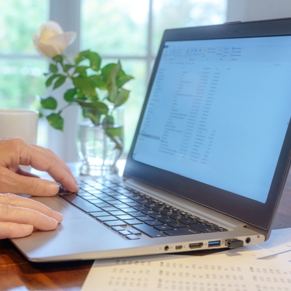 Hände einer älteren Person, die auf einem Laptop tippt. Im Hintergrund stehen eine Tasse und eine weiße Blume auf dem Schreibtisch. Hände einer älteren Person, die auf einem Laptop tippt. Im Hintergrund stehen eine Tasse und eine weiße Blume auf dem Schreibtisch.